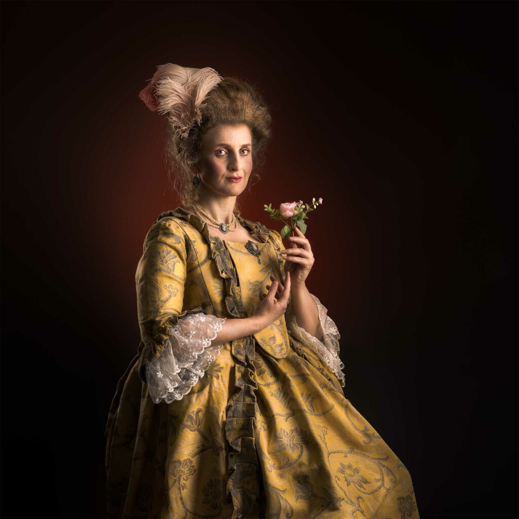 Portrait historique style XVIIIe siècle, femme en robe dorée faite main et perruque poudrée tenant une fleur, mise en scène en studio.
