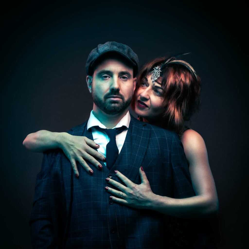 Portrait de couple inspiré de Peaky Blinders, homme en casquette et costume, femme l'enlaçant, séance couple créative en studio.