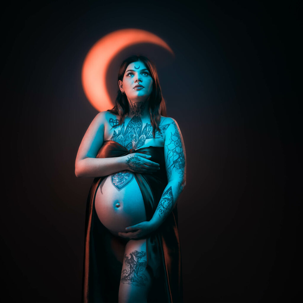 Photo de grossesse créative en studio, future maman tatouée devant une lune orange, par Éric Pothier à Rueil-Malmaison.