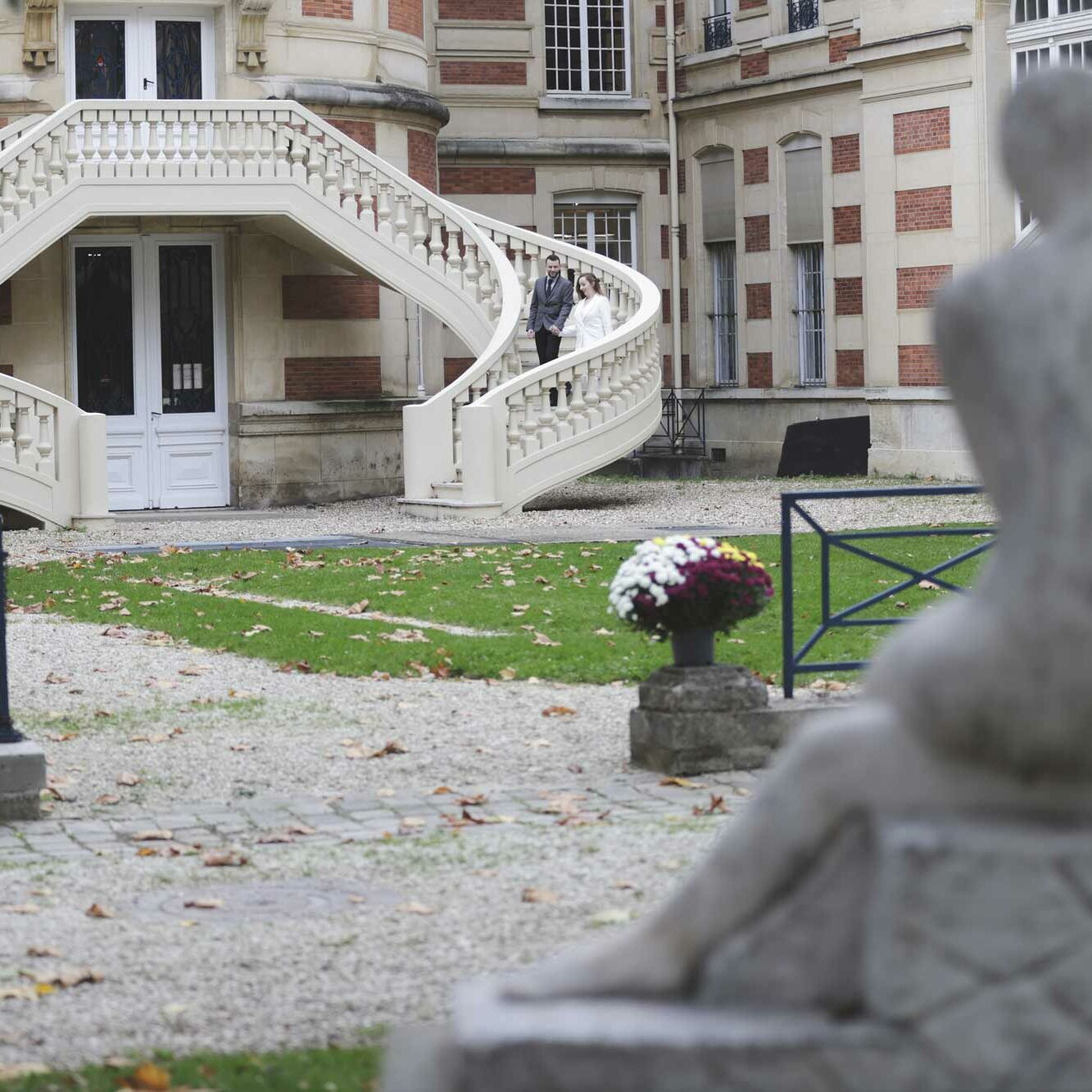 Mariés en haut de l’escalier en fer à cheval de l’hôtel de ville de Versailles, vus depuis le parc avec une statue au premier plan